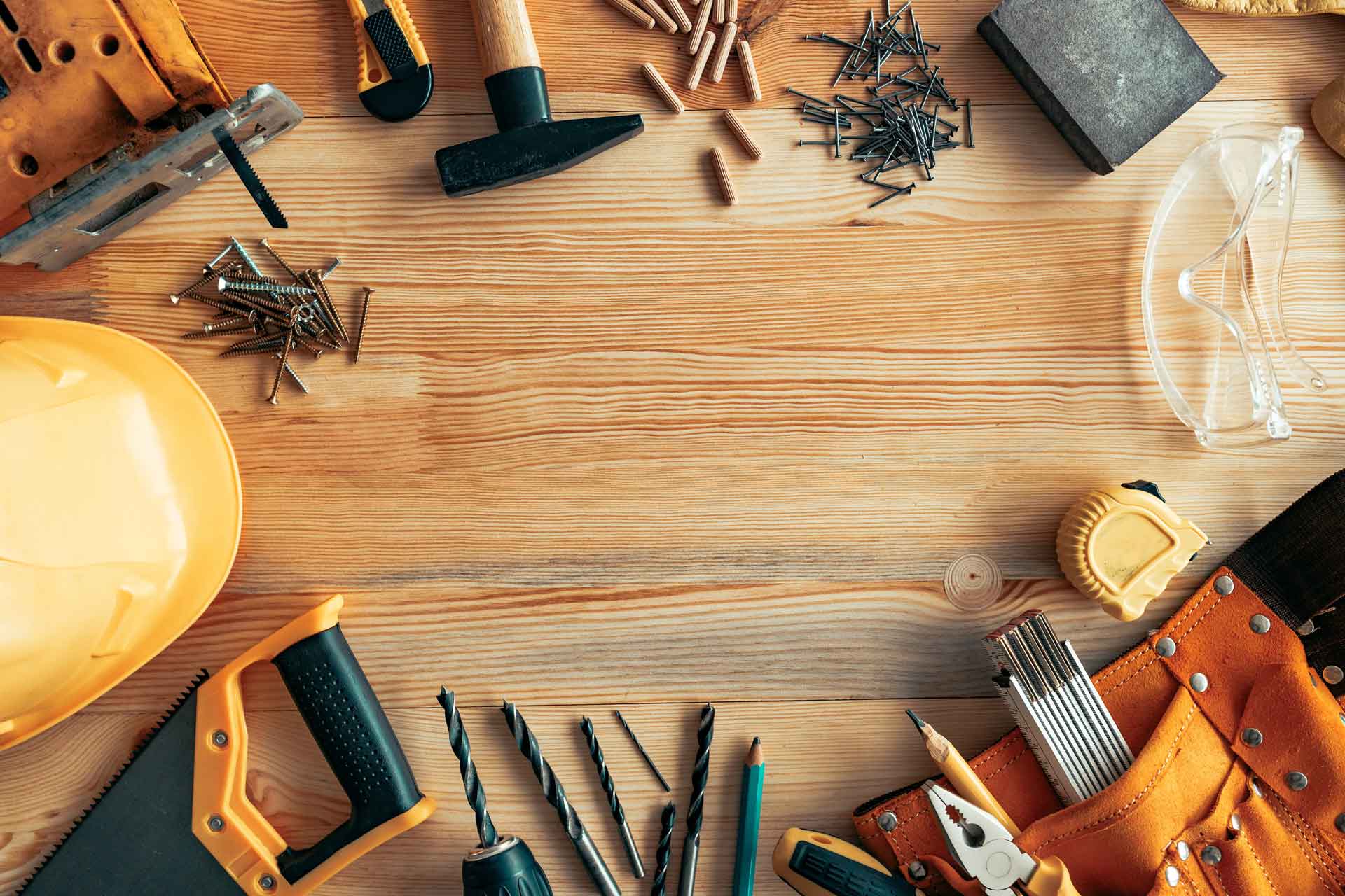 Wooden bench with an assortment of tools and construction supplies scattered across it: a hardhat, safety glasses, nails and screws, drill bits, a tape measure.